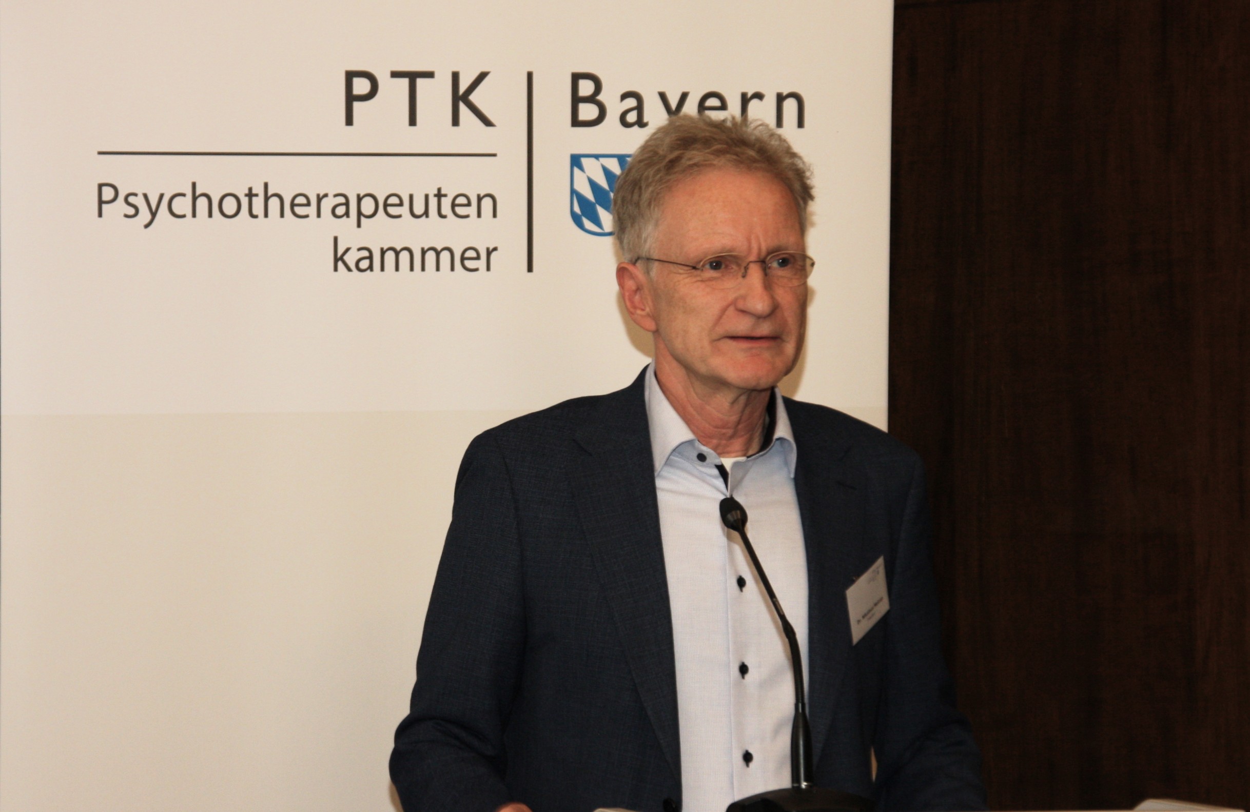 Ein Mann in einem dunkelblauen Anzug steht am Redepult. Im Hintergund ist ein Banner der PTK Bayern zu sehen.